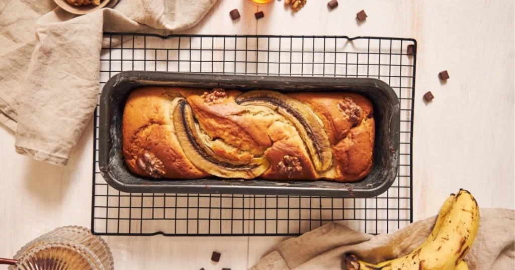 Freshly baked banana bread with a golden-brown crust, sliced and served on a wooden cutting board, surrounded by ripe bananas and ingredients used in the recipe