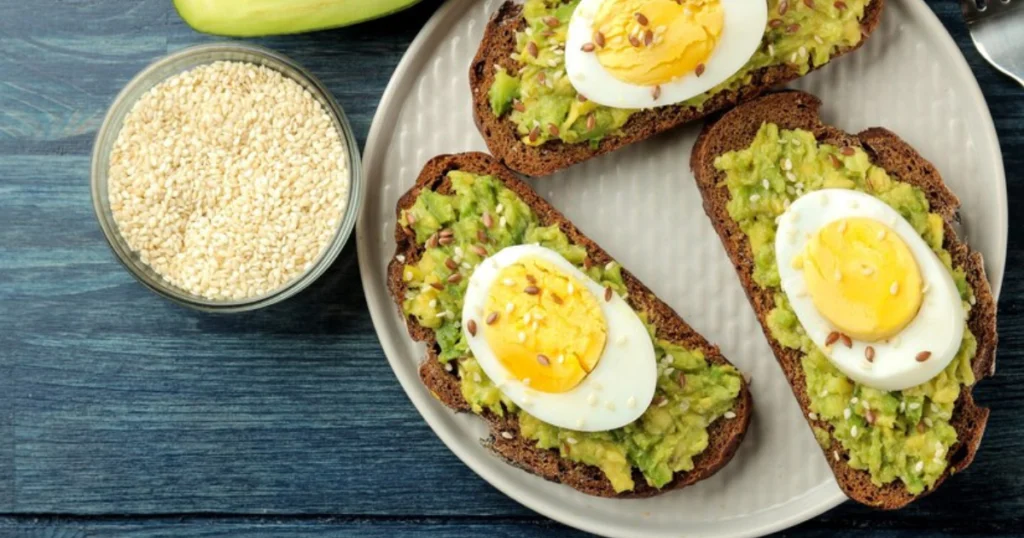 Avocado toast topped with sliced hard-boiled eggs on a rustic brown bread, garnished with sesame and flax seeds, served on a white plate alongside a bowl of sesame seeds.