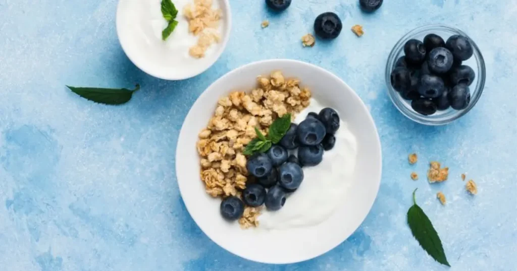 A bowl of Greek yogurt topped with granola, fresh blueberries, and a mint sprig on a bright blue background